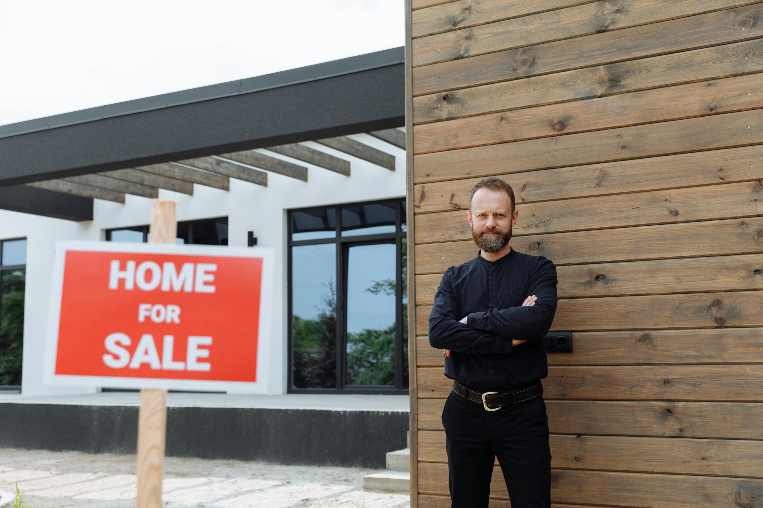 Real estate agent standing by a modern home with a 'Home for Sale' sign.